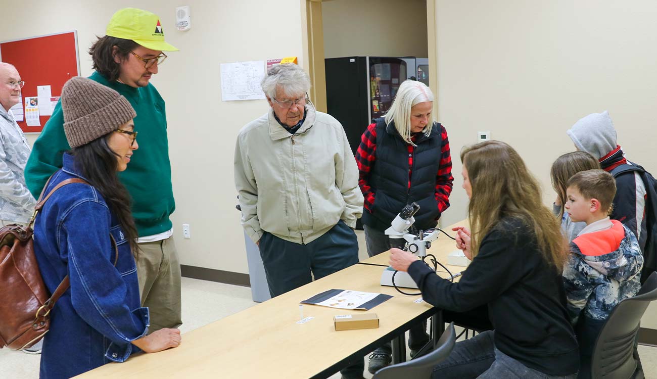 Community members of different ages stand around instructor at a microscope