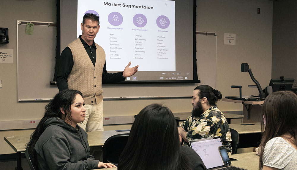 Male instructor stands in front of white board talking to students