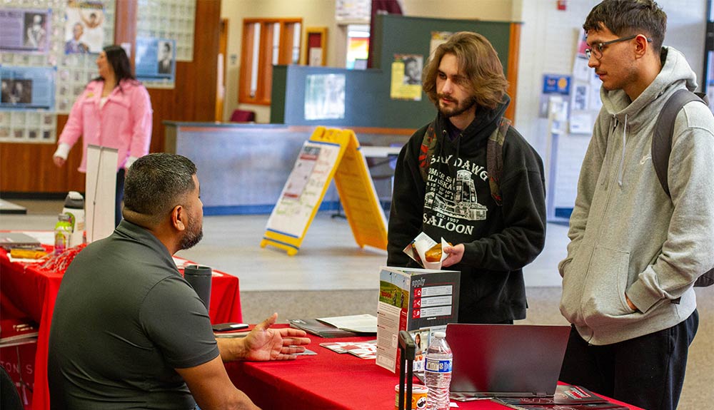 Two male students talk to man sitting at table