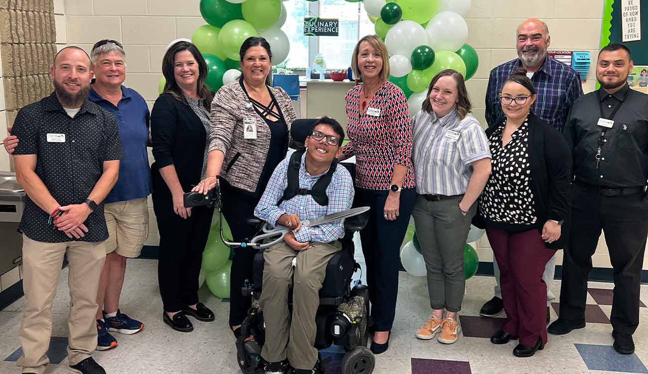 Student in wheelchair hold scissors with other people standing to sides at ribbon cutting ceremony