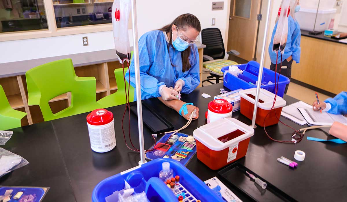 A phlebotomy student takes blood from a sim arm during lab