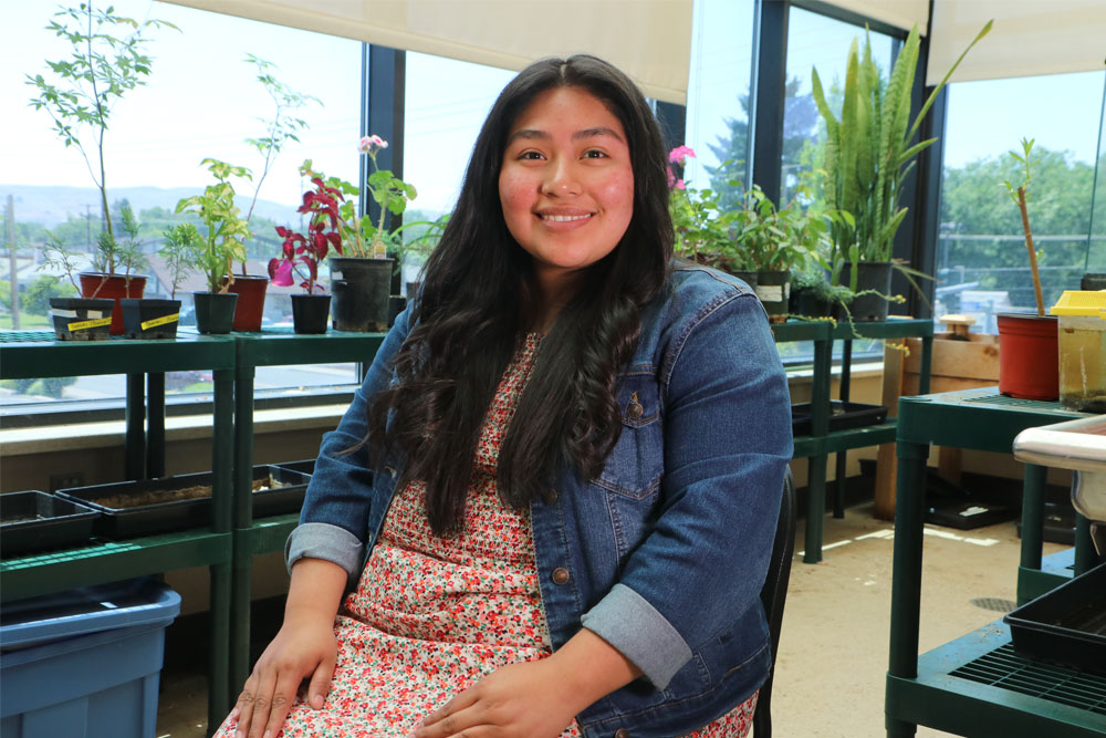 Annamaria Delaluz poses for Student Story picture sitting in a chair with potted plants in the background. 