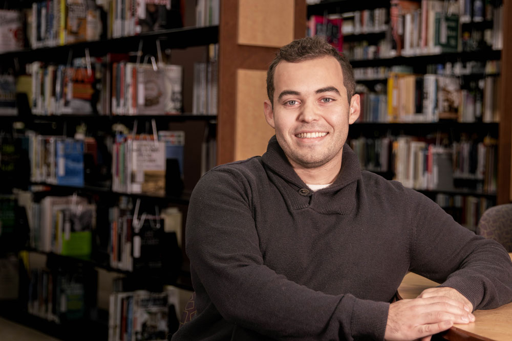 Caleb Valdez-Copeland poses for Student Story picture in the YVC Library with book shelves in the background. 