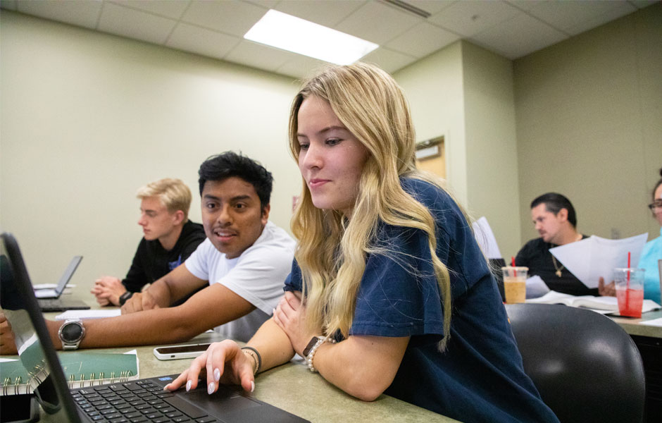 Student works on a computer in class while another student looks on. 