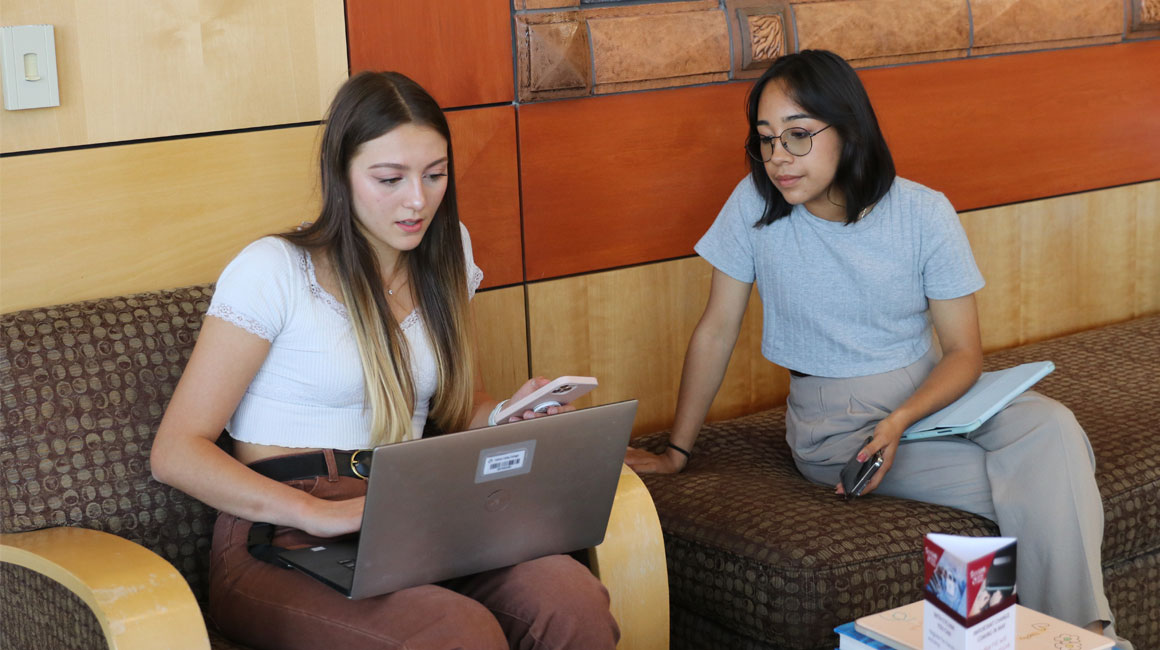 Two students sitting in a common area working on a lap top. 